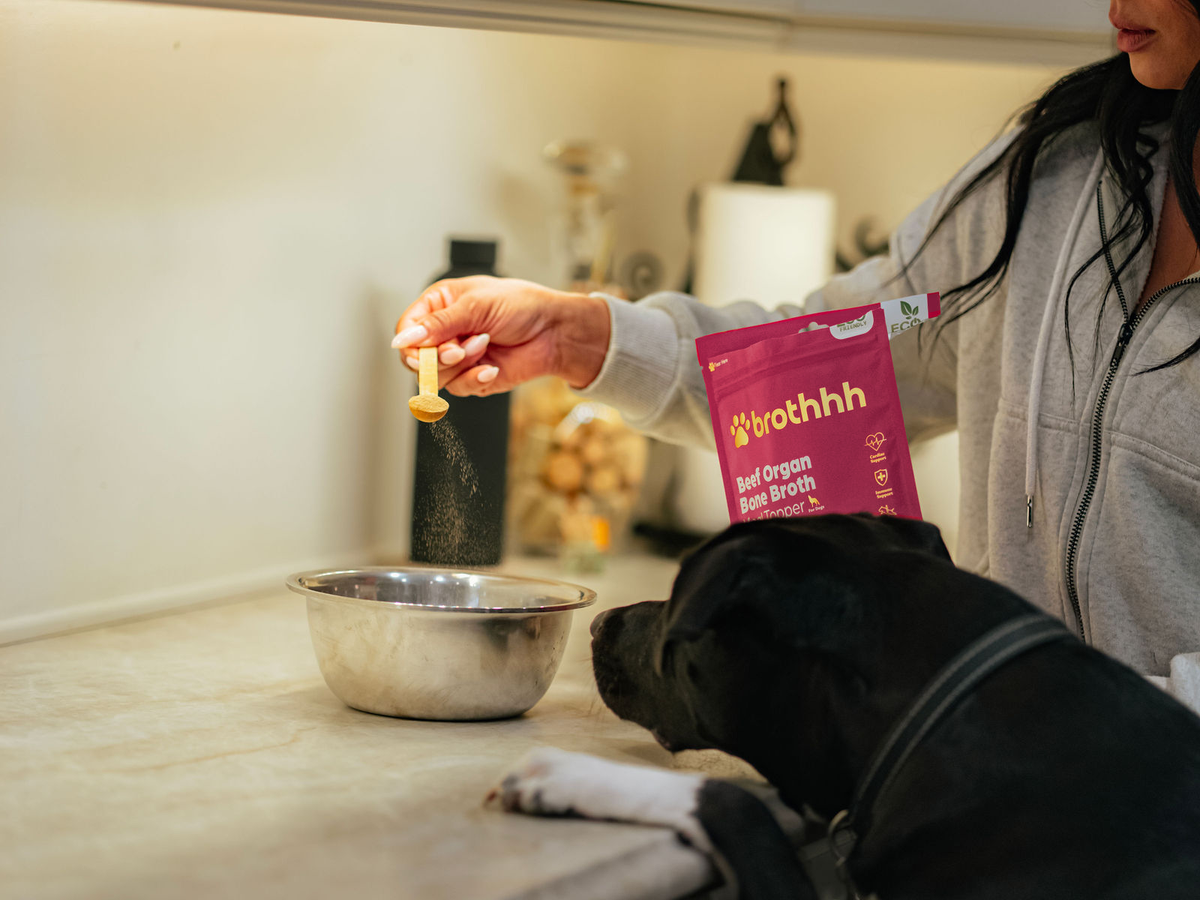 A woman preparing The Meal Topper by Brothhh with her dog beside her