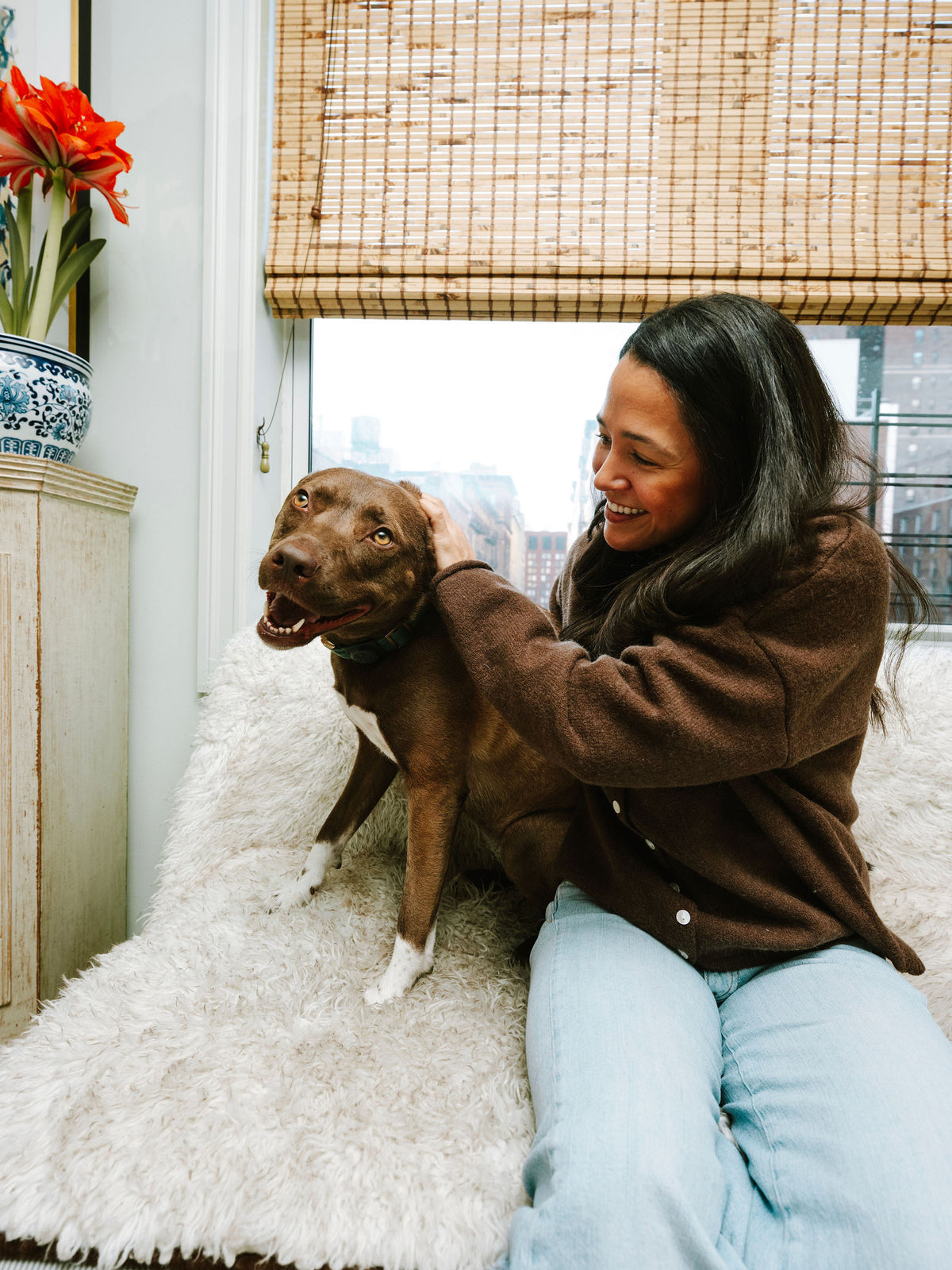 A woman smiling as she pets her brown dog on a cream chair at home