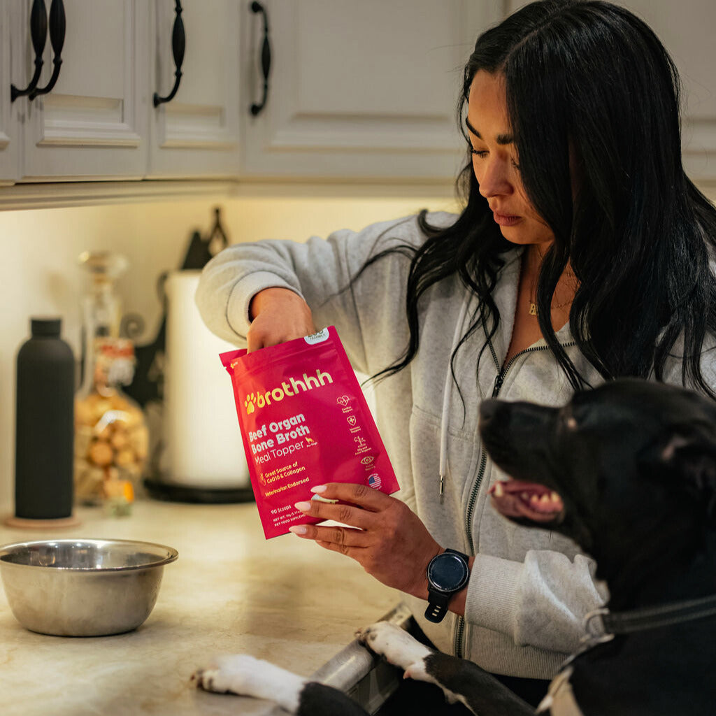 A woman preparing The Meal Topper by Brothhh with her dog beside her in the kitchen